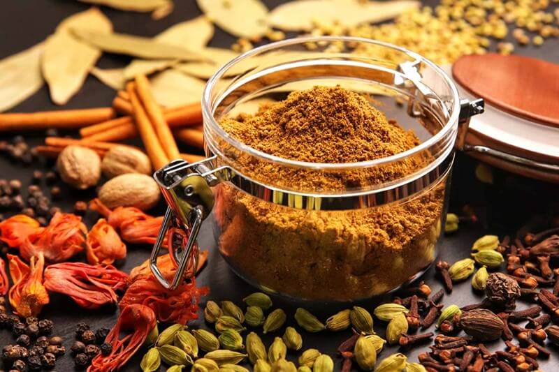 Garam masala đựng trong hũ kín, xung quanh là các loại gia vị nguyên liệu như tiêu, thảo quả, hoa hồi, nhục đậu khấu